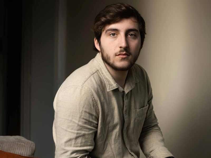 Portrait d'un jeune homme aux cheveux courts et barbe légère, portant une chemise claire, assis dans une pièce lumineuse.