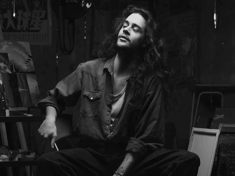 Homme aux cheveux longs et chemise ouverte, assis dans un atelier artistique, regardant vers la gauche dans une ambiance sombre.