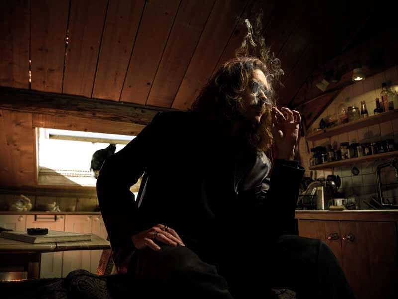 Homme aux cheveux longs fumant une cigarette dans une pièce en bois, avec une lumière tamisée et une atmosphère intime.