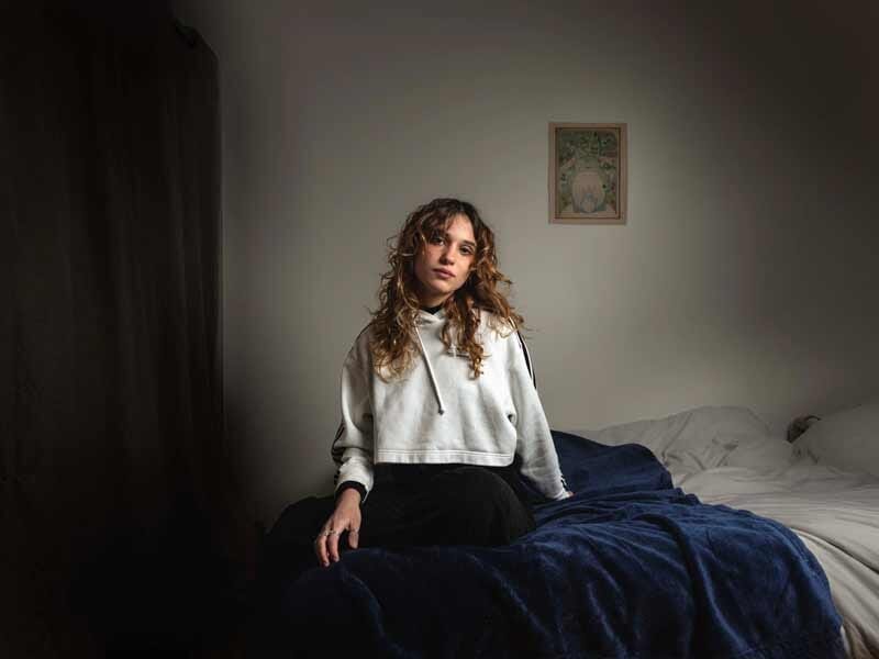 Jeune femme assise sur un lit avec une couverture bleue, portant un sweat blanc, dans une chambre éclairée naturellement.