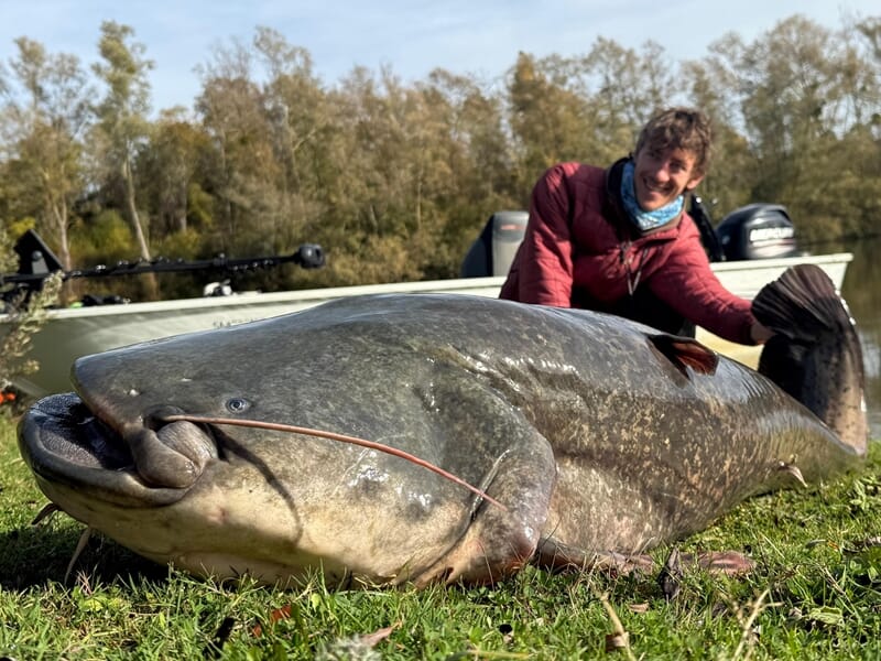 Homme souriant assis sur l'herbe près d'un très gros poisson lors d'un stage de pêche en bateau sur la Seine à Poissy.