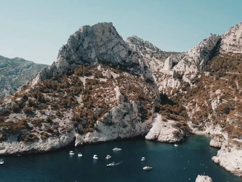 Vue aérienne d'une calanque avec plusieurs petits bateaux ancrés dans une eau bleue entourée de falaises rocheuses.