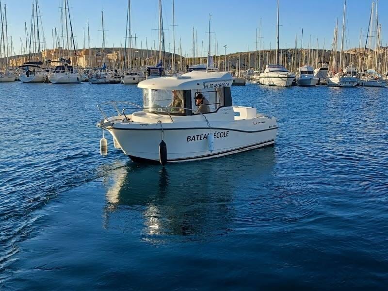 Petit bateau-école blanc naviguant sur une mer calme avec un port de plaisance et de nombreux voiliers en arrière-plan.