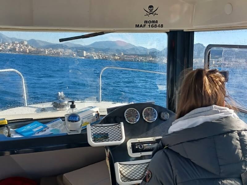 Personne vue de dos à la barre d'un bateau, avec tableau de bord et vue sur la mer et la côte en arrière-plan, passant le permis bateau à Marseille.