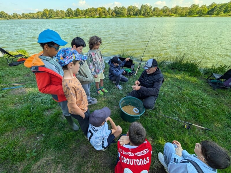 Un groupe d'enfants attentifs écoute un moniteur de pêche près d'un lac, lors d'un stage de pêche à Vichy.