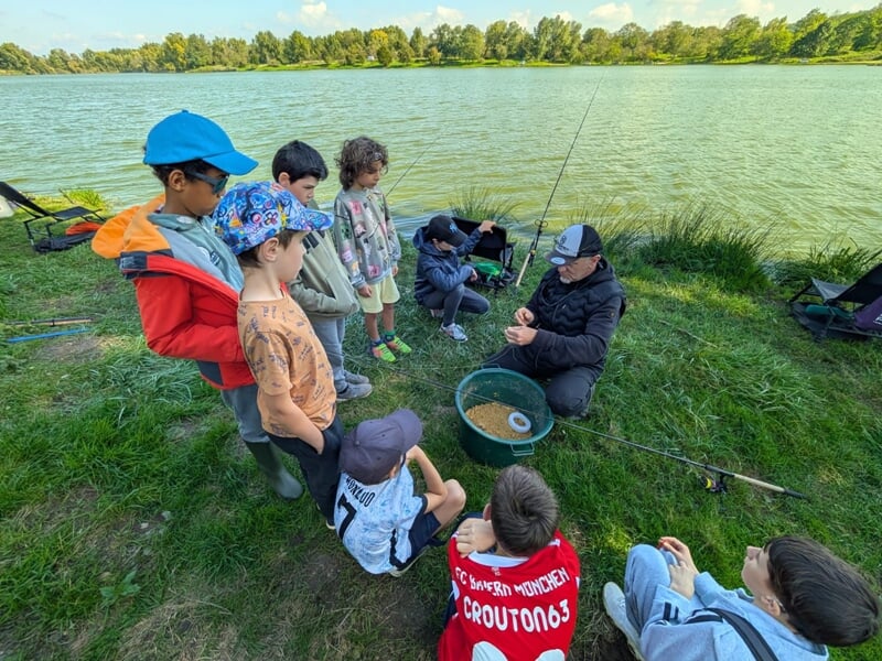 Un groupe d'enfants attentifs écoute un moniteur de pêche près d'un lac, lors d'un stage de pêche.