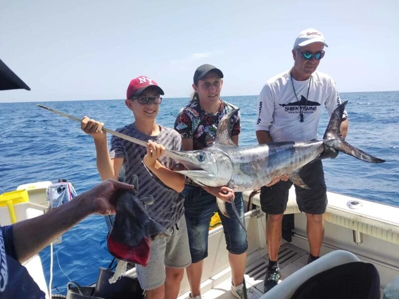 Trois personnes souriantes sur un bateau exhibant un poisson long et argenté lors d'une sortie pêche à Saintes-Maries-de-la-Mer.