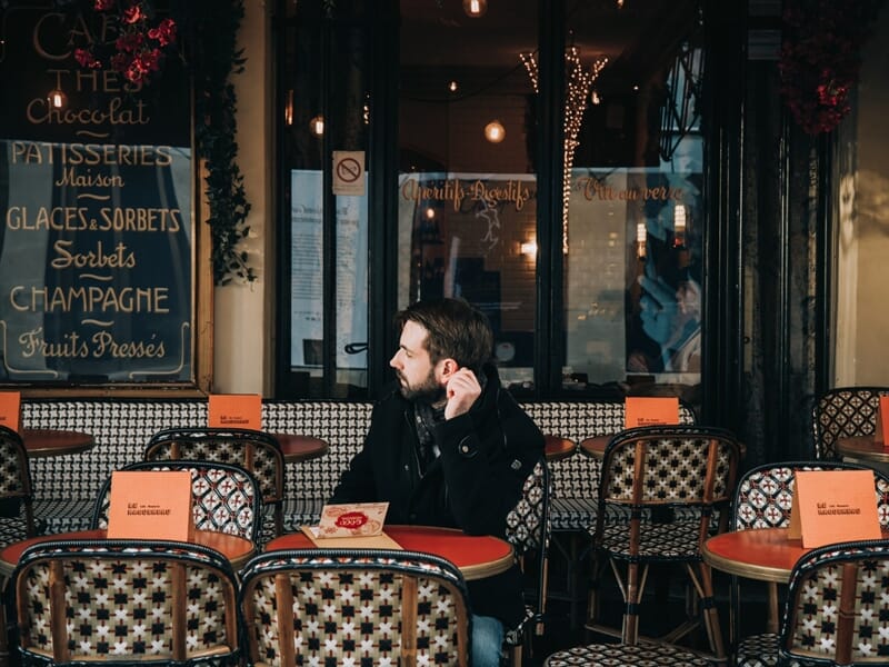 Homme assis seul à une table de café parisien, regardant vers la gauche lors d'un shooting photo lifestyle à Maisons-Laffitte