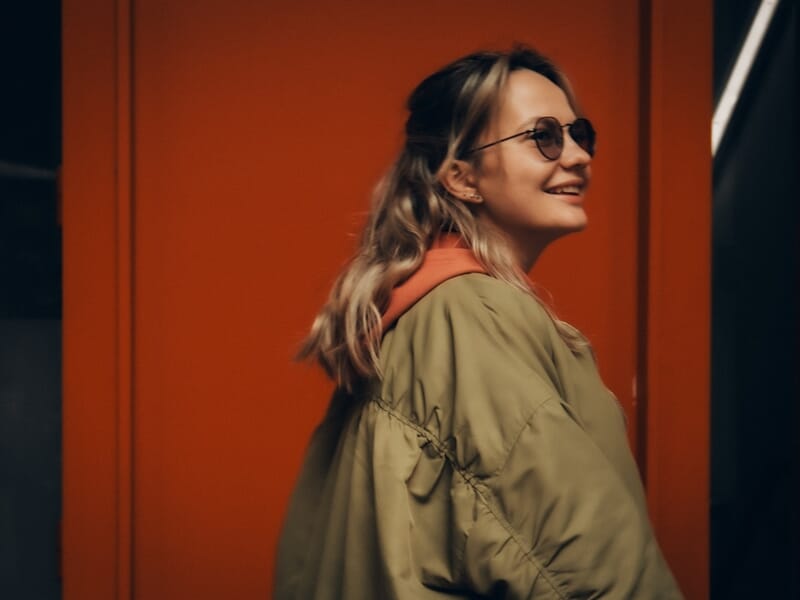 Jeune femme souriante portant des lunettes de soleil et un manteau kaki lors d'un shooting photo lifestyle à Maisons-Laffitte.