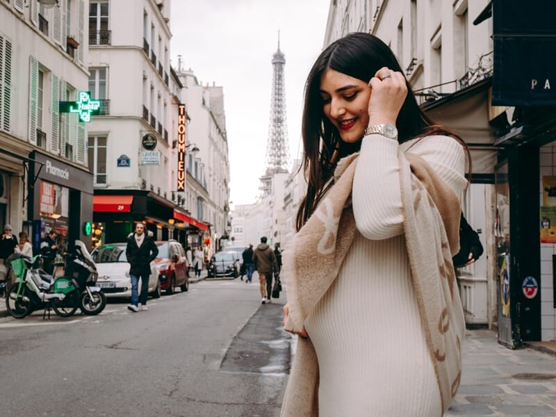 Jeune femme souriante en manteau beige dans une rue animée avec la tour Eiffel lors shooting photo grossesse à Maisons-Laffitte. 
