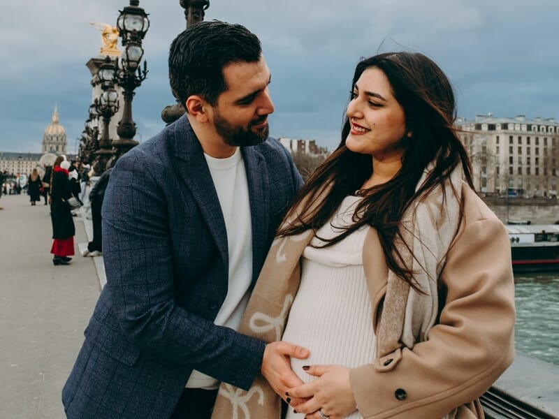 Couple heureux avec femme enceinte, homme touchant son ventre, sur un pont parisien lors shooting photo grossesse à Maisons-Laffitte. 
