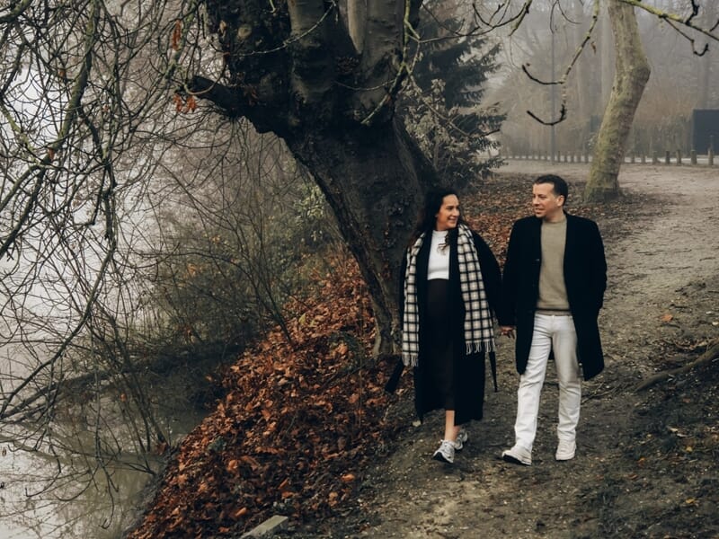 Couple marchant main dans la main dans un parc d'automne avec arbres dénudés et feuilles mortes lors shooting photo grossesse à Maisons-Laffitte. 