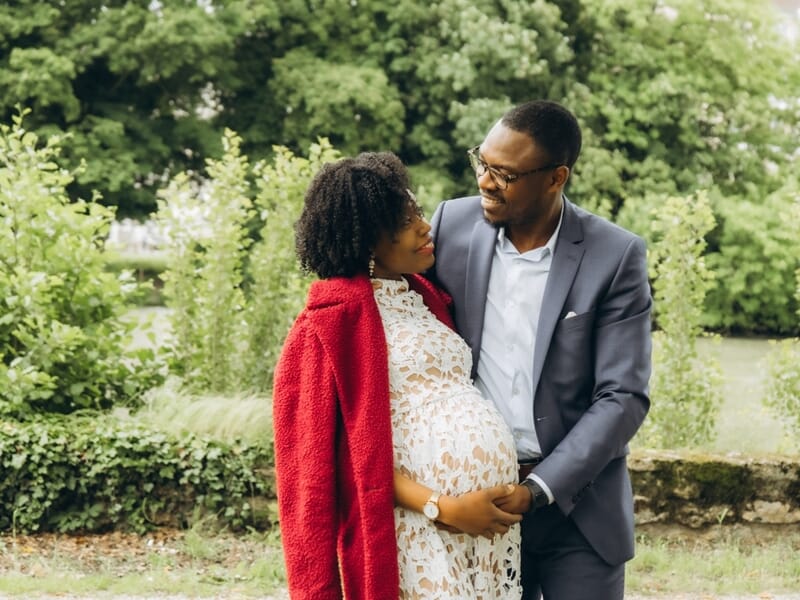 Couple souriant dans un jardin verdoyant, femme enceinte en robe blanche et manteau rouge lors shooting photo grossesse à Maisons-Laffitte. 
