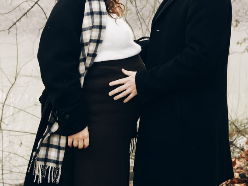 Couple en manteaux noirs et écharpe, homme posant la main sur le ventre lors shooting photo grossesse à Maisons-Laffitte. 