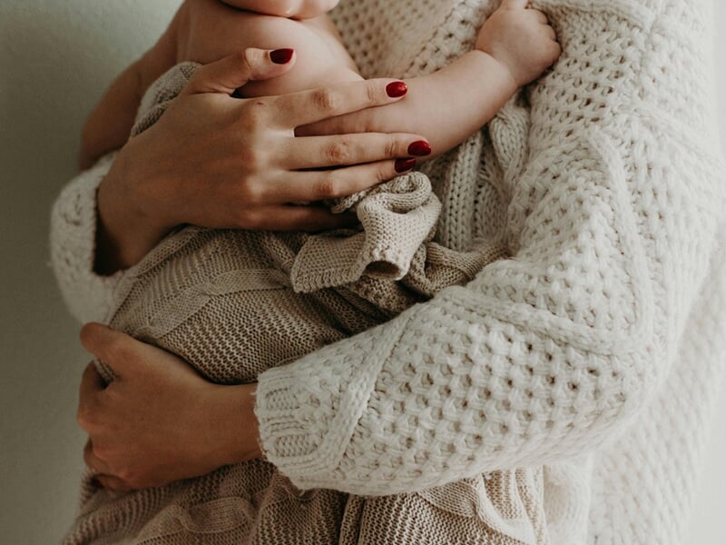 Gros plan sur une mère en pull tricoté tenant son bébé enveloppé dans une couverture lors d'un shooting avec un photographe bébé à Maisons-Laffitte.