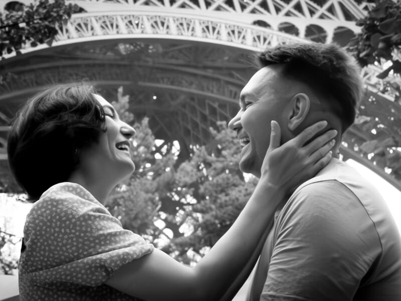 Un couple souriant se regarde tendrement sous la tour Eiffel, photo en noir et blanc lors d'un shooting photo couple à Maisons-Laffitte.