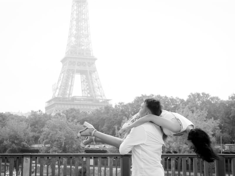 Un homme porte une femme dans ses bras sur un pont avec la tour Eiffel en arrière-plan lors d'un shooting photo couple à Maisons-Laffitte.