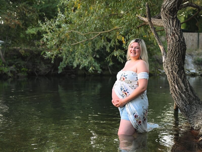 Femme enceinte souriante debout dans l'eau d'une rivière, entourée d'arbres verts en arrière-plan.