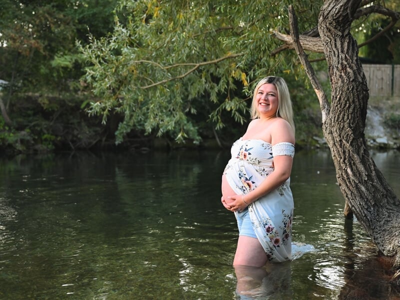 Femme enceinte souriante debout dans l'eau d'une rivière, entourée d'arbres verts en arrière-plan.