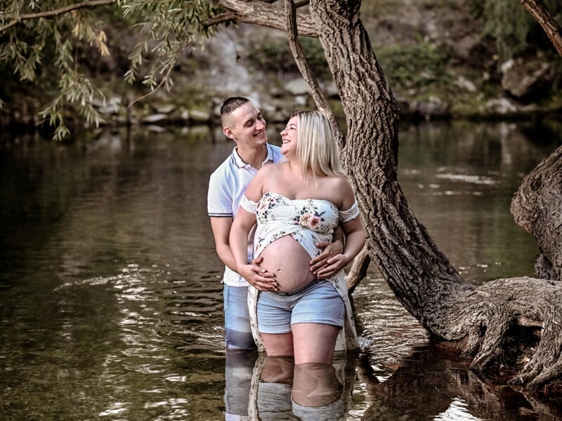Couple heureux dans l'eau d'une rivière, l'homme embrasse la femme enceinte qui porte une robe fleurie.
