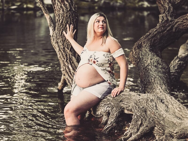 Femme enceinte assise sur une branche d'arbre au bord de l'eau, regardant vers le haut avec un air rêveur.