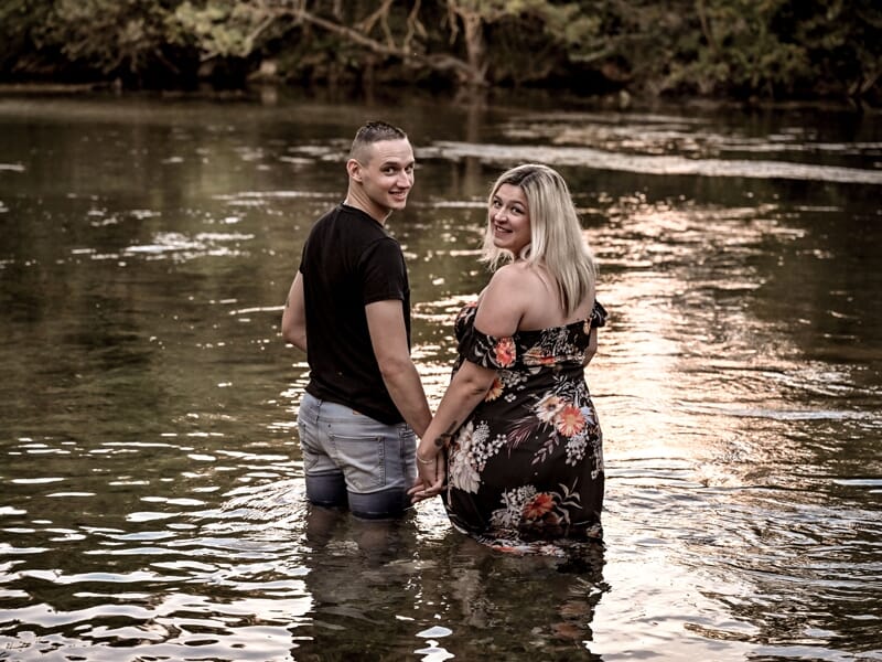 Couple main dans la main dans l'eau d'une rivière, la femme porte une robe noire à fleurs, tous deux souriants.
