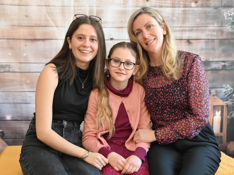 Trois femmes souriantes assises sur un canapé devant un mur en bois, posant pour une photo de famille chaleureuse.