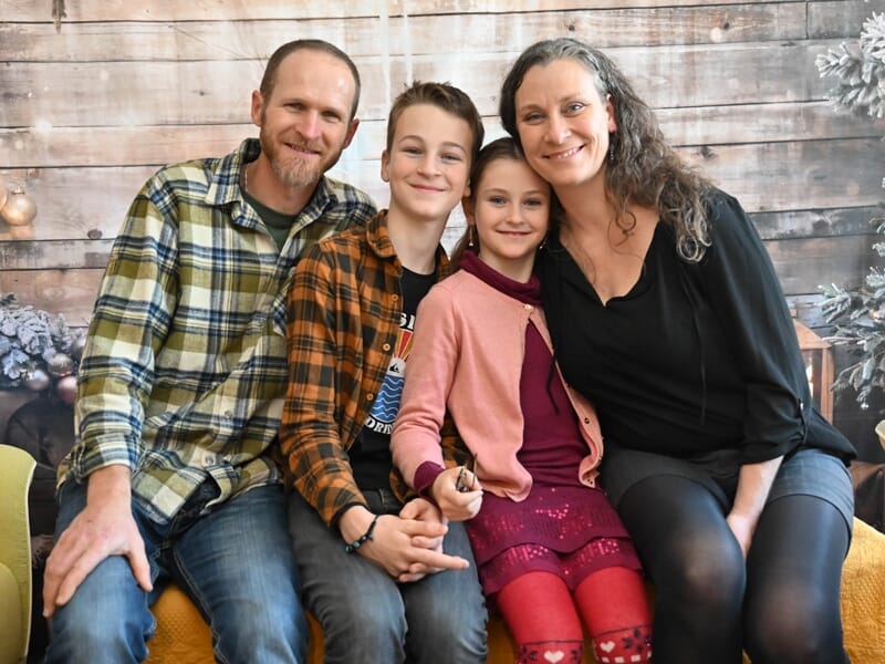 Quatre personnes souriantes, deux adultes et deux enfants, assises sur un canapé avec un décor en bois en arrière-plan.