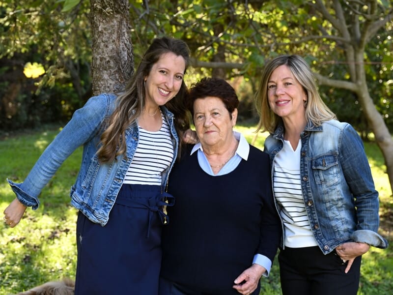 Trois femmes de différentes générations posent ensemble dans un jardin ensoleillé, souriantes et heureuses.