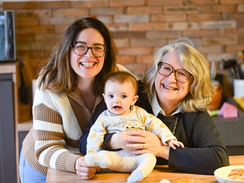 Deux femmes et un bébé souriants assis à une table en bois dans une cuisine chaleureuse et accueillante.