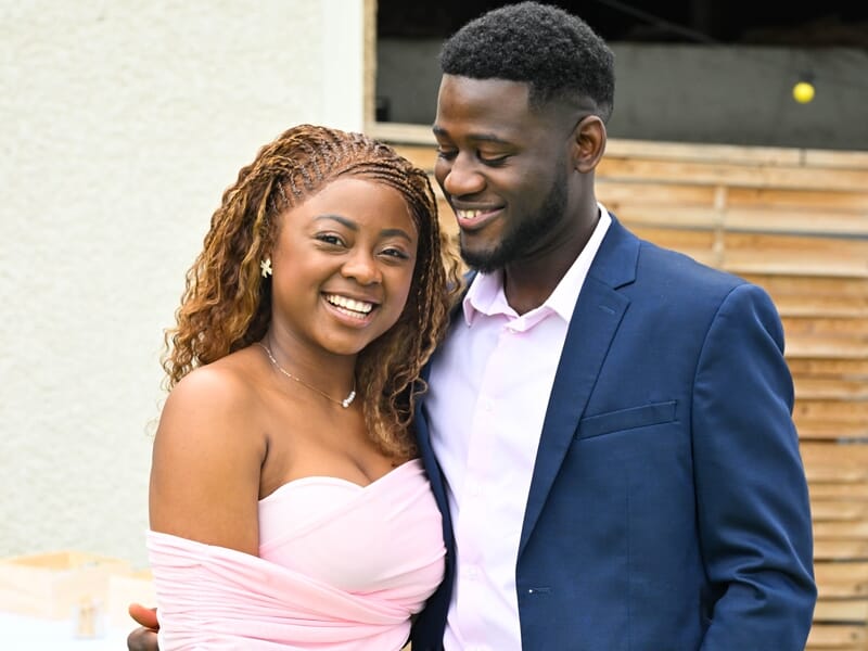 Jeune couple souriant, femme en robe rose et homme en costume bleu, ambiance joyeuse en extérieur.
