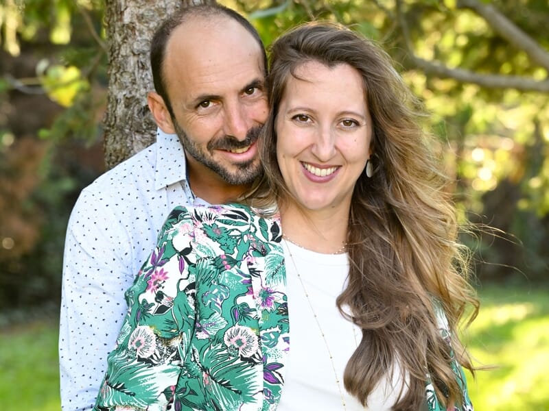 Couple heureux posant près d’un arbre, femme en veste fleurie et homme en chemise blanche à pois.