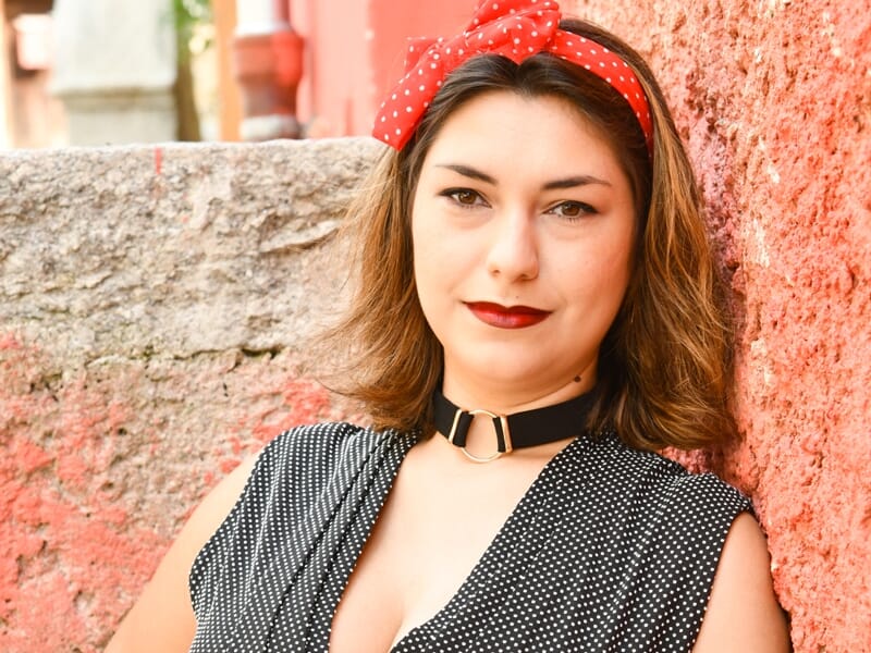 Portrait d'une jeune femme aux cheveux bruns avec un bandeau rouge à pois blancs, posant devant un mur rose et pierre.