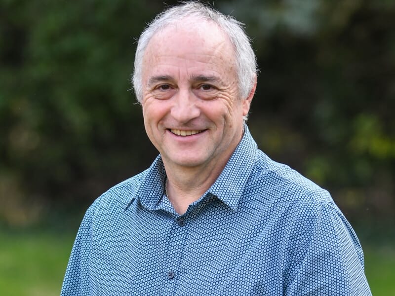 Homme souriant d'âge mûr portant une chemise bleue à motifs, debout dans un jardin avec un fond flou de verdure.