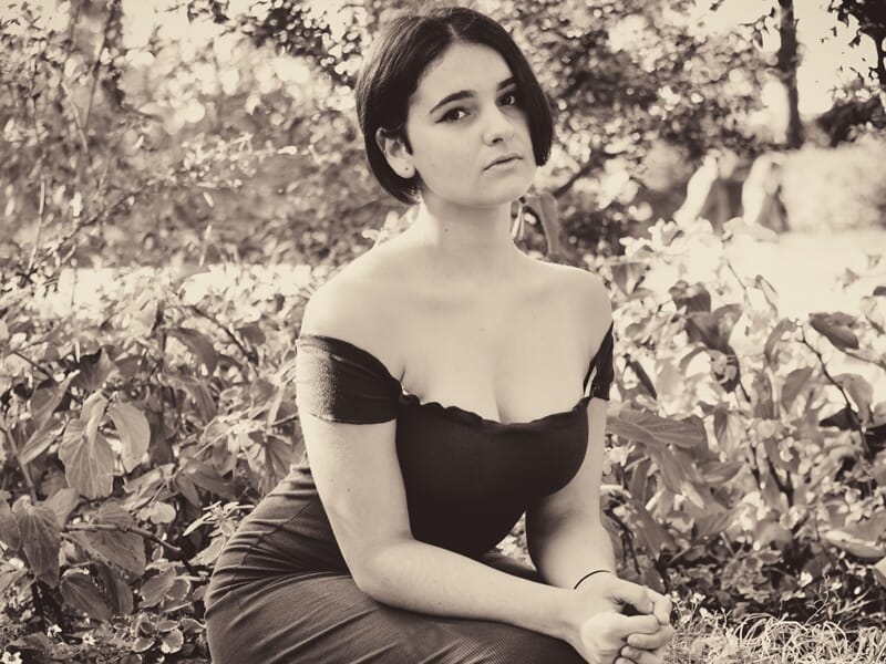 Jeune femme en robe noire sans bretelles, assise dans un jardin, photo en noir et blanc avec un style vintage.