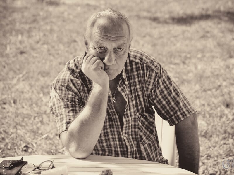 Homme d'âge mûr assis à une table extérieure, appuyant sa tête sur sa main, regard pensif et sérieux.