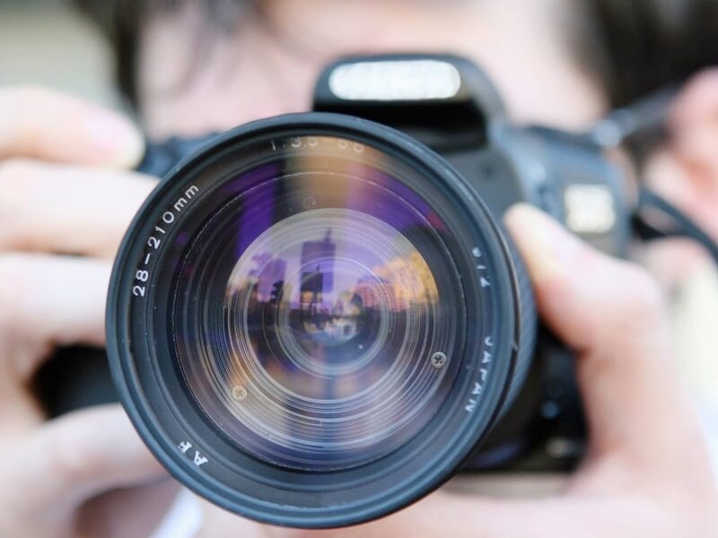Gros plan sur l'objectif d'un appareil photo reflex tenu par une personne en arrière-plan floue.