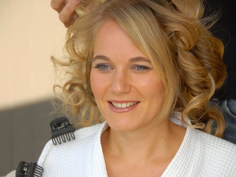 Femme souriante aux cheveux bouclés en train de se faire coiffer, portant une robe blanche.