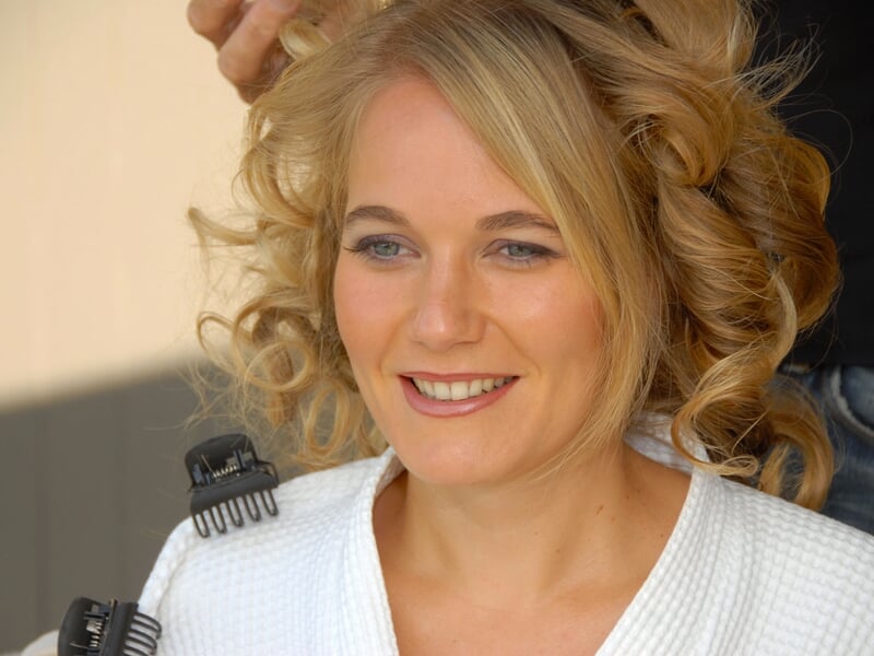 Femme souriante aux cheveux bouclés en train de se faire coiffer, portant une robe blanche.