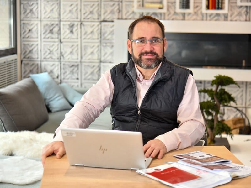 Homme souriant avec lunettes et gilet noir, assis à une table en bois avec un ordinateur portable et des documents, intérieur moderne.