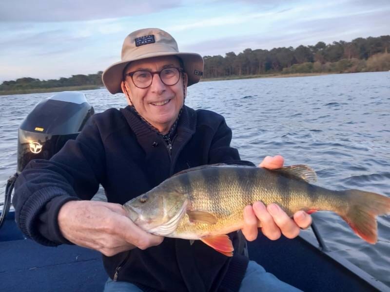 Homme souriant tenant un poisson jaune rayé sur un bateau au milieu d'un lac calme sous un ciel clair.