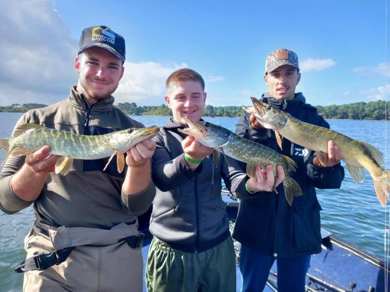 Trois jeunes hommes debout sur un bateau, chacun tenant un grand brochet fraîchement pêché, lors d'un stage de pêche à Dax.
