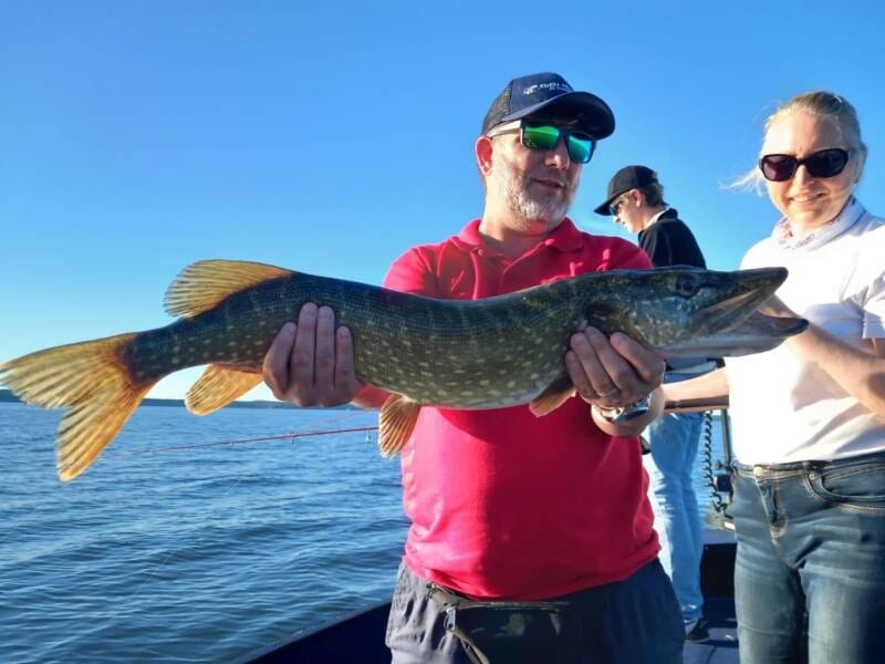 Homme en t-shirt rouge tenant un gros brochet sur un bateau, une femme souriante à côté de lui.