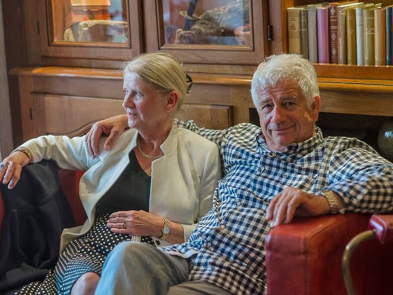 Un couple de personnes âgées assises sur un canapé rouge dans une pièce avec une bibliothèque en arrière-plan.