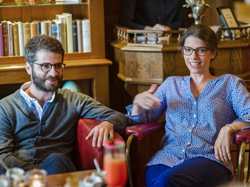 Un homme et une femme souriants assis dans un salon avec des meubles en bois et des livres derrière eux.