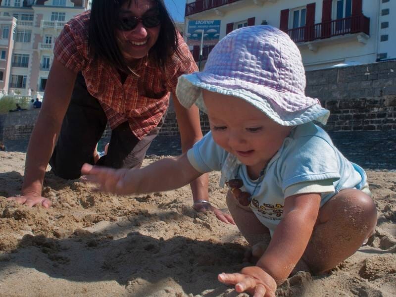 Lors d'un shooting photo à Biarritz, un bébé joue dans le sable sur une plage, sous la surveillance attentive d'une femme souriante en arrière-plan.