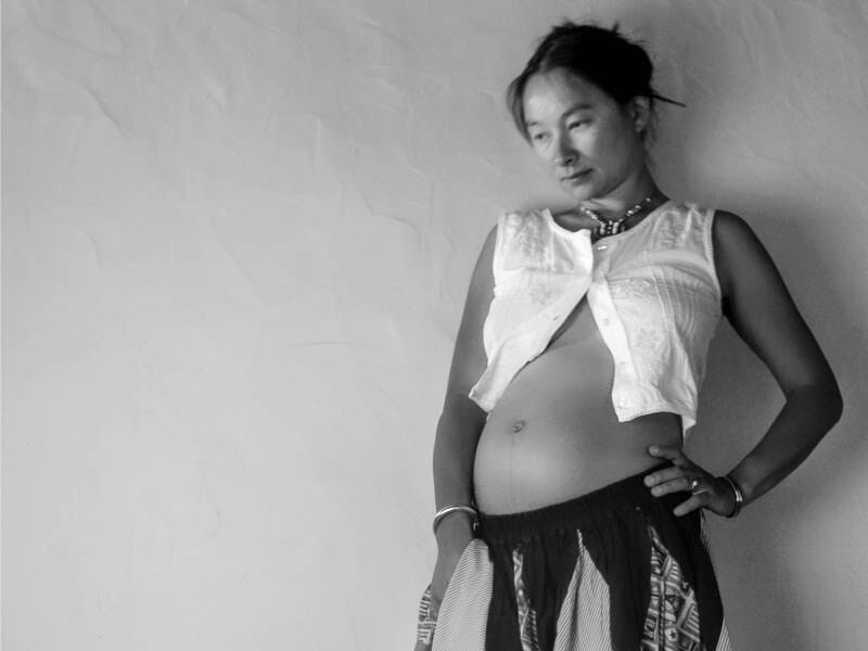 Femme enceinte posant devant un mur clair, vêtue d'un gilet blanc et d'une jupe colorée, le regard pensif, lors d'un shooting photo à Biarritz.