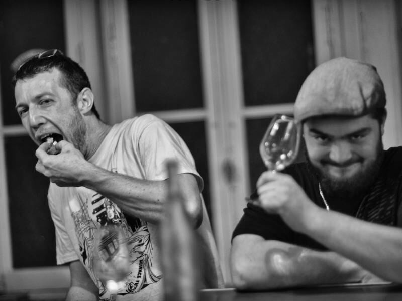 Deux hommes à table, l'un mange tandis que l'autre tient un verre de vin en souriant, ambiance détendue et amicale.