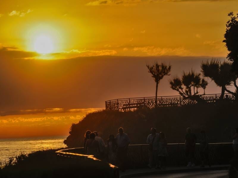 Coucher de soleil sur la côte de Biarritz lors d'un stage photo, avec silhouettes de personnes marchant le long d'une promenade en bord de mer.