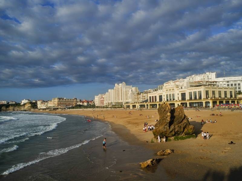 Plage de sable avec quelques baigneurs et bâtiments en arrière-plan sous un ciel nuageux.